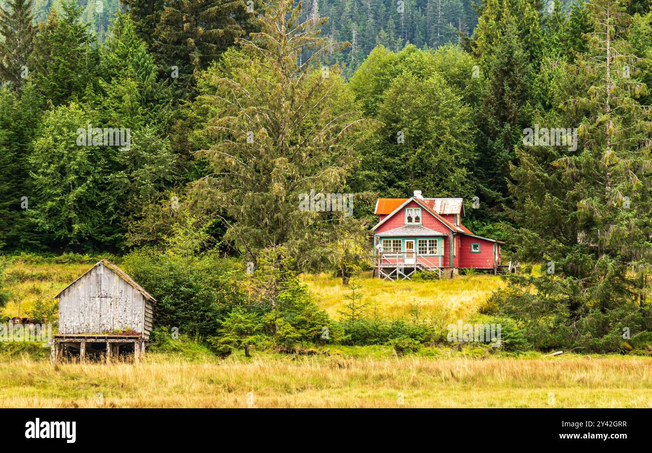 Altes rotes Haus in einem Feld, umgeben von großen Bäumen, mit einem kleineren Bootsschuppen in der Nähe. Nordküste von BC auf Porcher Island. Stockfoto
