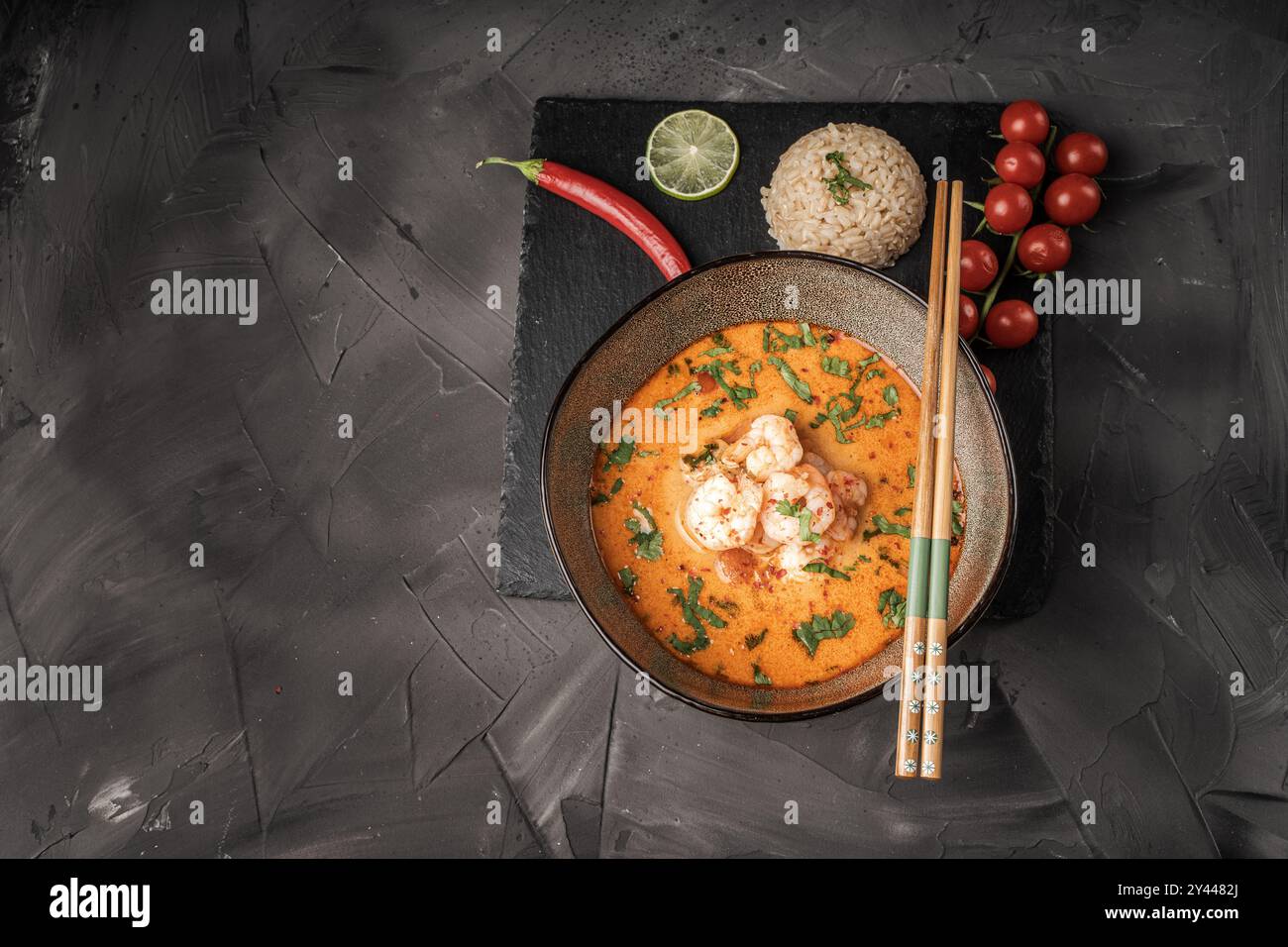 Tom Yum Kung scharfe Thai-Suppe mit Garnelen in einer Schüssel auf dunklem Hintergrund und rotem Pfeffer, Reis und Limette mit Koriander und Essstäbchen auf einer schwarzen Glimmerboa Stockfoto