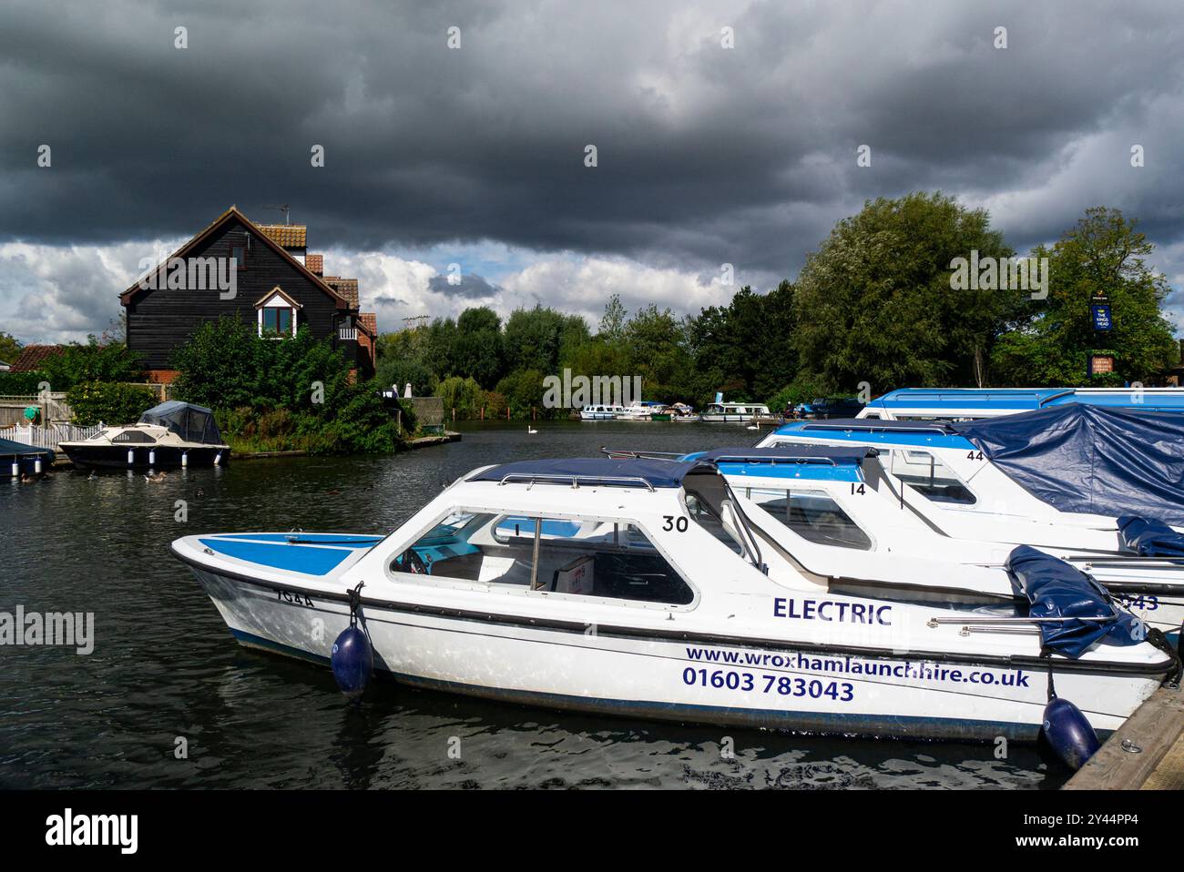 Blick entlang des Flusses Bure im Dorf Wroxham in den Norfolk Broads, bekannt als Hauptstadt der Broads, mit vertäuten Elektromotorbooten zum Verleih von NOR Stockfoto