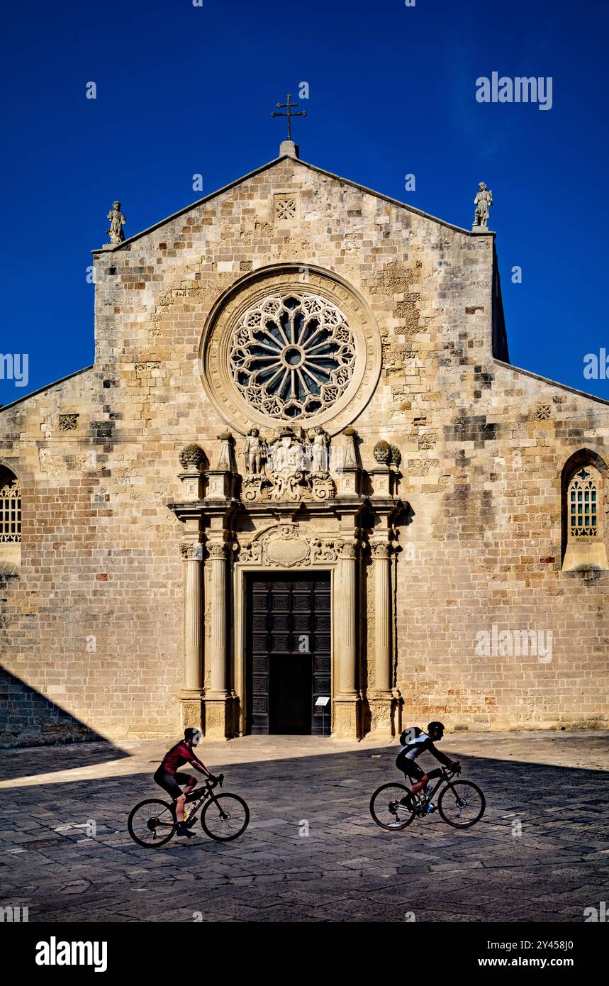 Zwei Radfahrer radeln an der Vorderseite der romanischen Kathedrale von Otranto vorbei. Die katholische Basilika stammt aus dem 11. Jahrhundert und befindet sich in Otranto, Apulien, Stockfoto