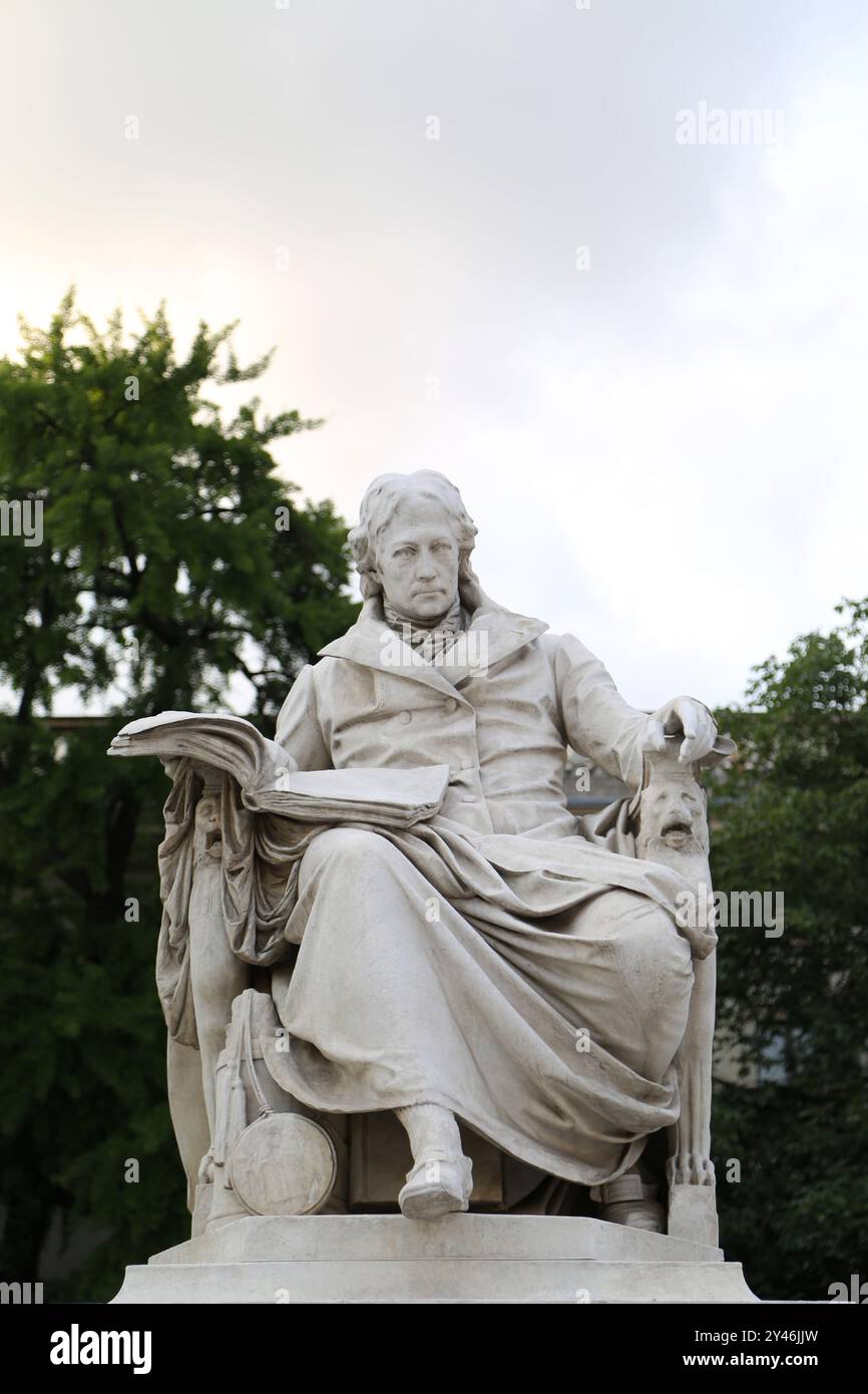 Die Statue von Wilhelm von Humboldt sitzt anmutig vor seiner gleichnamigen Universität in Berlin, umgeben von üppigem Grün, und symbolisiert seine Profession Stockfoto