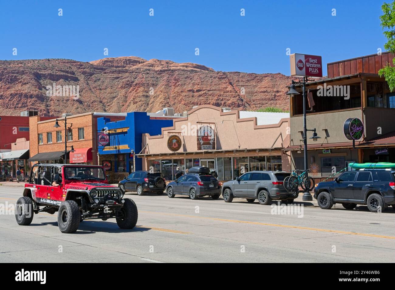 Geländefahrzeug, das die Main Street mit rotem Hintergrund der Moab-Felgen-Klippen entlang fährt – Moab Utah, April 2024 Stockfoto