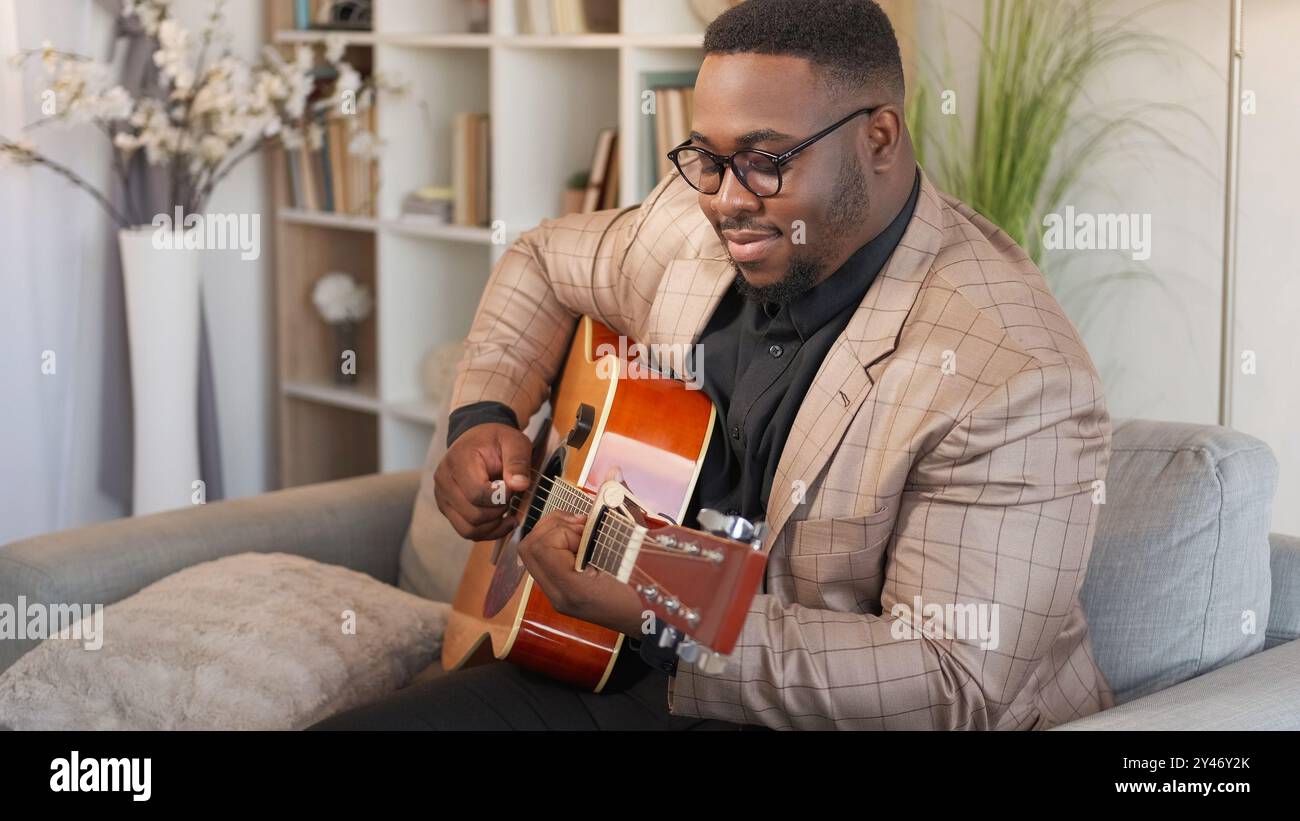 Gitarrenspieler. Kreatives Hobby. Talentierter, inspirierter, lächelnder Mann Musiker, der Melodie auf einem akustischen Streichinstrument auf der Couch im Wohnzimmer spielt. Stockfoto