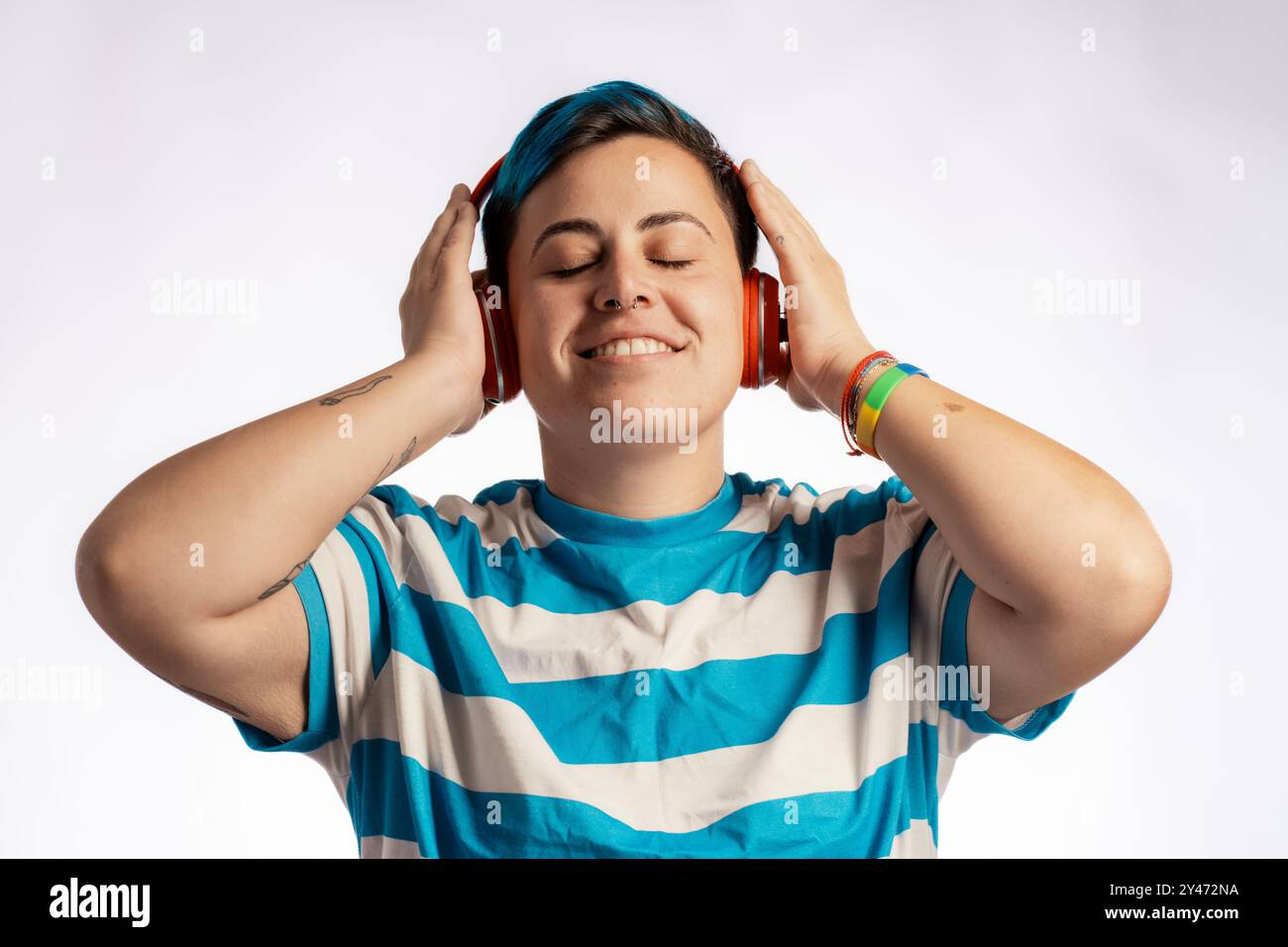 Glückliches Geschlecht, das sich nicht anpasst, mit blauem Haar und gestreiftem Hemd, das Musik mit roten Kopfhörern hört. Ausdrucksstarkes Porträt, das Freude zeigt, di Stockfoto