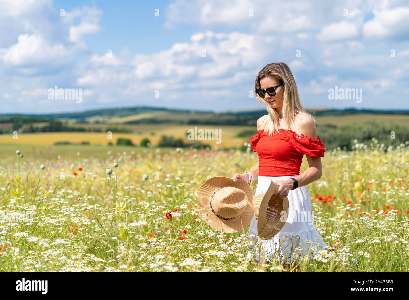 Blonde Frau in Sonnenbrille, rot-weißem Kleid auf einem wilden Blumenfeld, Strohhüte in den Händen Stockfoto