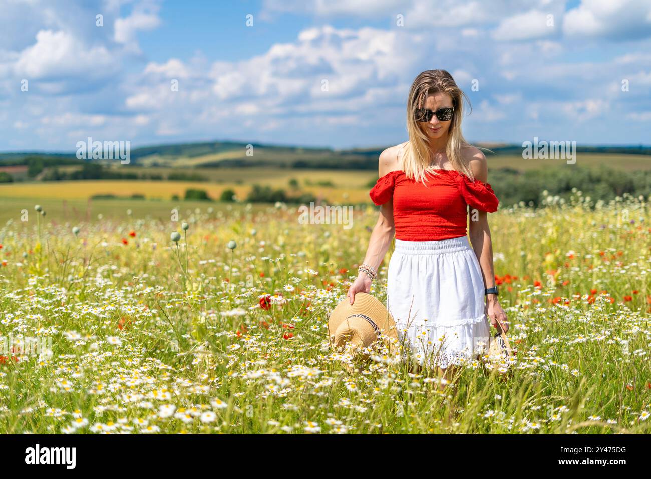 Blonde Frau in Sonnenbrille, rot-weißem Kleid auf einem wilden Blumenfeld, Strohhüte in den Händen Stockfoto
