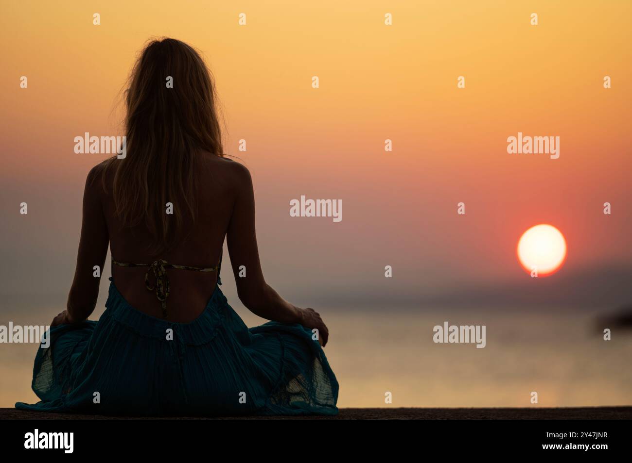 Blick von hinten auf ein junges Frauenkleid, das in Lotusposition am Meer bei schönem, warmem Sonnenaufgang sitzt. Sommerurlaub, Ruhe Stockfoto