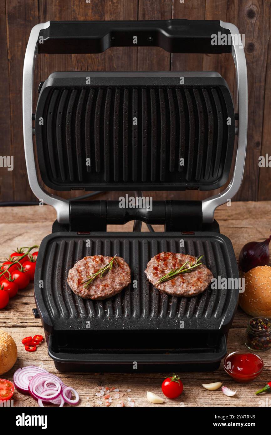 Rinderpasteten für einen Burger werden auf einem eleganten elektrischen Grill auf rustikalem Holzhintergrund gebraten. Hausgemachte Hamburger. Zutaten für Burger. Stockfoto