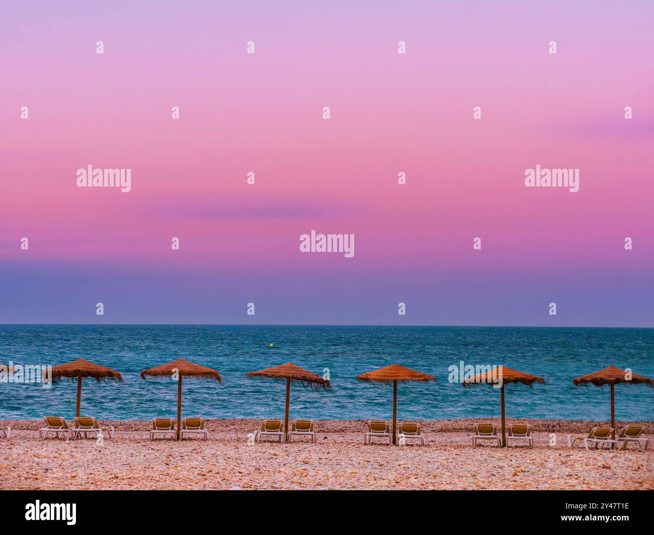 Eine ruhige Strandszene mit mehreren Strohschirmen, die an einem mit Kieselsteinen übersäten Ufer vor dem Sonnenuntergangshimmel aufgereiht sind, Altea, Spanien Stockfoto