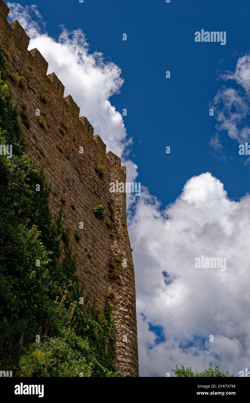 Eine Steinmauer der mittelalterlichen Burg Óbidos erhebt sich zu einem leuchtend blauen Himmel, der von flauschigen weißen Wolken gesäumt ist Stockfoto