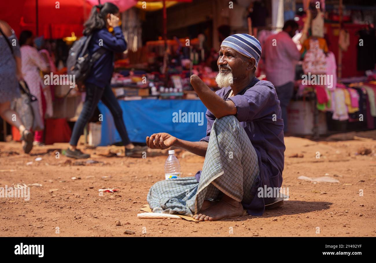 Mapusa, Indien - 19. Februar 2024: Ein alter Mann sitzt mit amputiertem Arm auf dem Boden und bittet um Almosen. Stockfoto