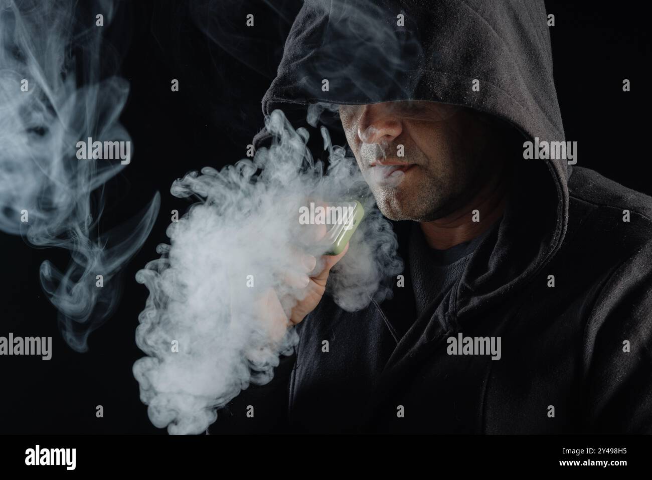 Mann in der Haube, der Dampf ausatmet, umgeben von rauchendem Rauch. Dunkler Hintergrund, elektronisches Zigarettenkonzept Stockfoto