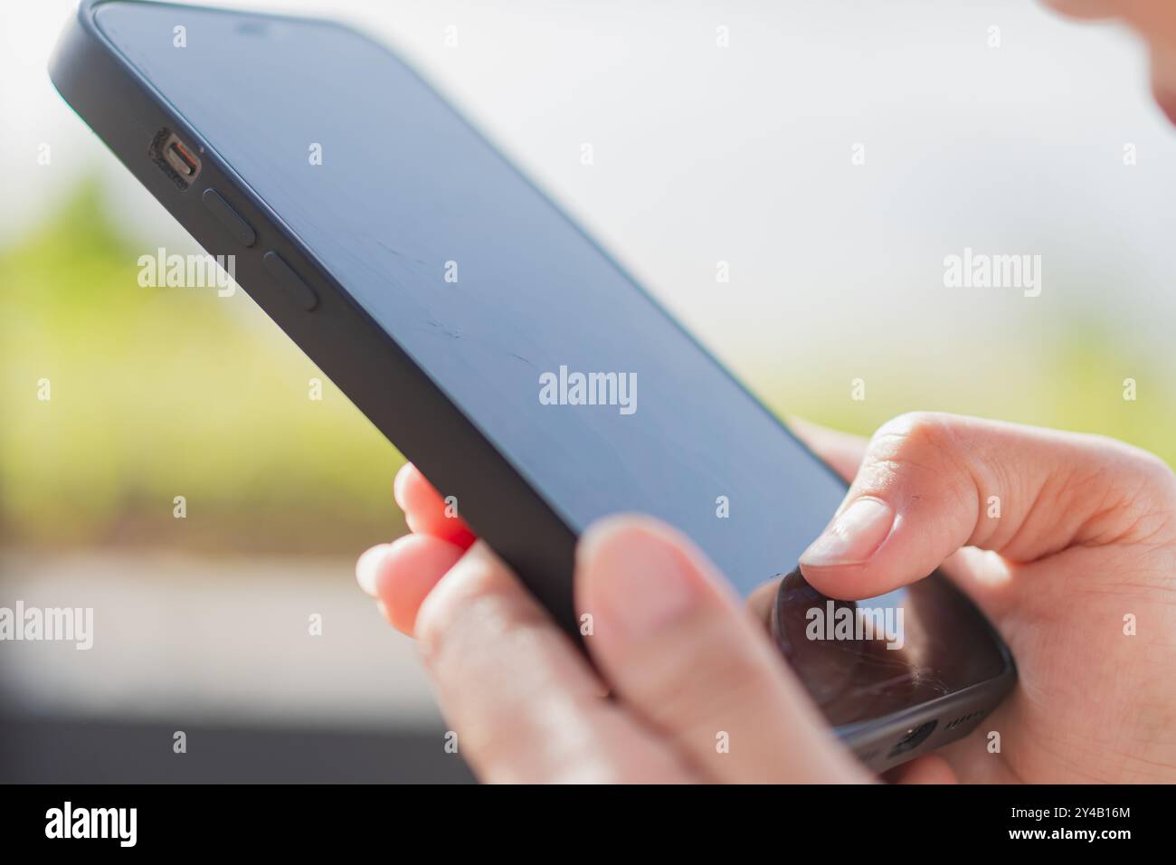 Eine Nahaufnahme einer Person, die ein Smartphone hält und sich auf den Bildschirm konzentriert. Der Hintergrund ist verschwommen, was auf eine grüne Umgebung im Freien hindeutet. Die Pers Stockfoto