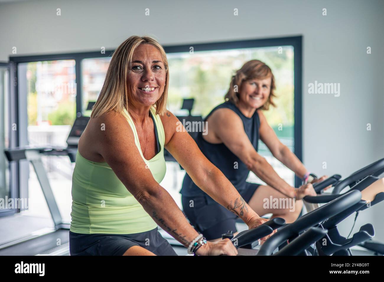 Zwei ältere sportliche Frauen lächeln in die Kamera, während sie im Fitnessstudio an einem Spinning-Kurs auf Fahrrädern teilnehmen. Das Bild zeigt, wie viel Spaß sie haben Stockfoto