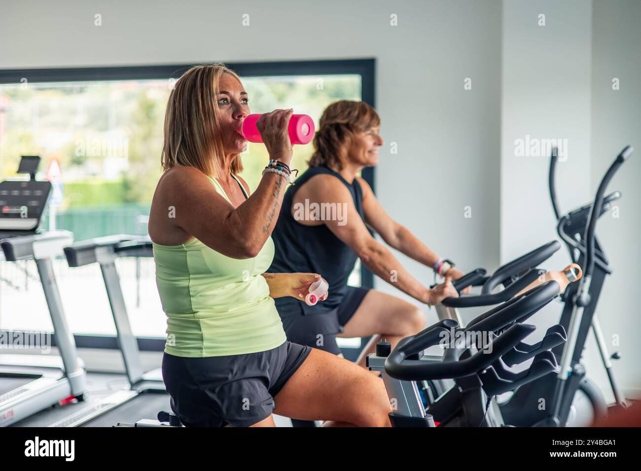 Eine reife sportliche Frau trinkt während eines Spinning-Kurses im Fitnessstudio aus einer rosafarbenen Wasserflasche. Das Bild zeigt, wie sie eine Pause einlegt und Hydrate hält Stockfoto