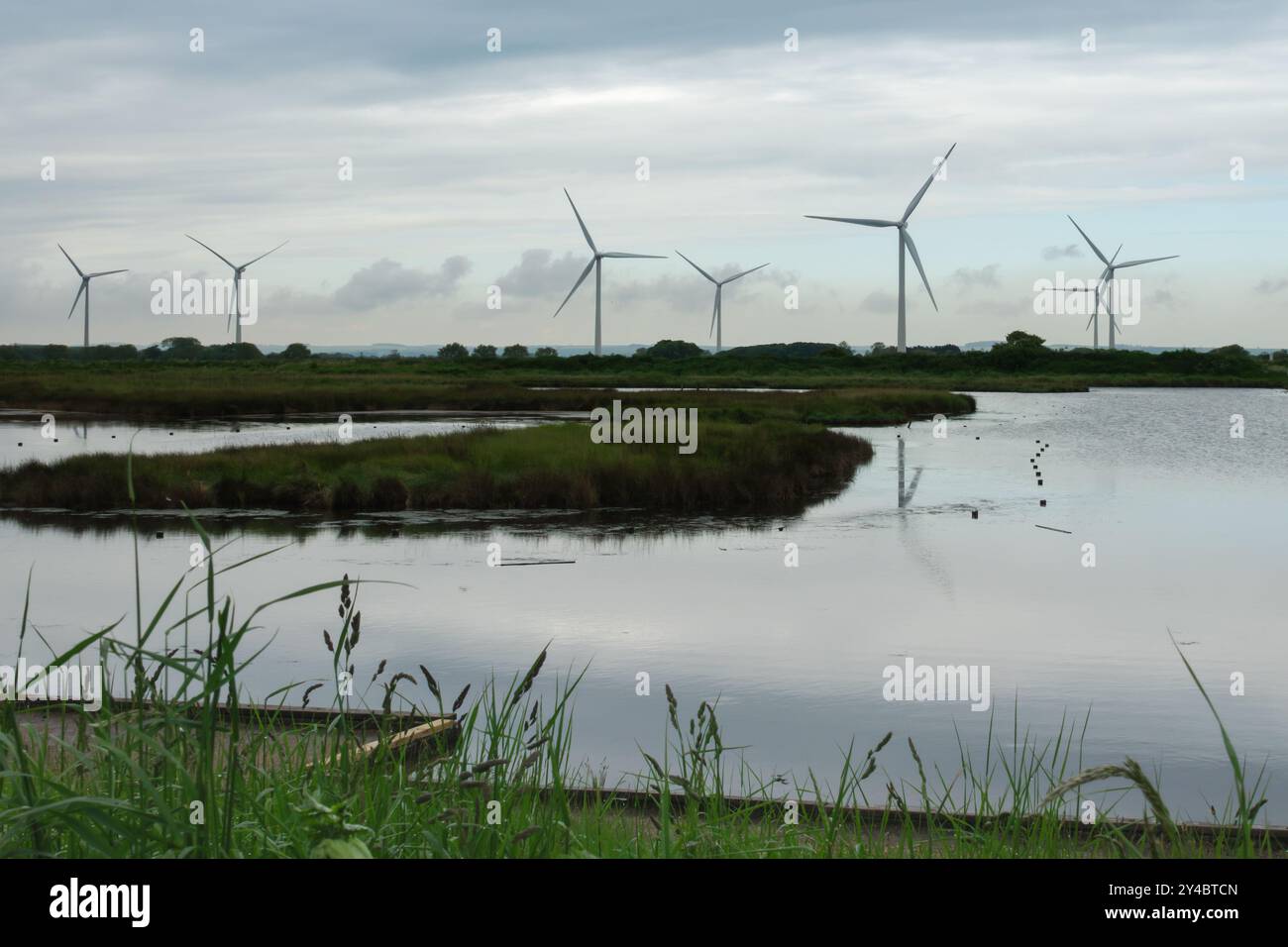 Eine Reihe von Windturbinen steht hoch vor einem bewölkten Himmel, ihre Reflexionen spiegeln sich in einem stillen, ruhigen Gewässer. Stockfoto