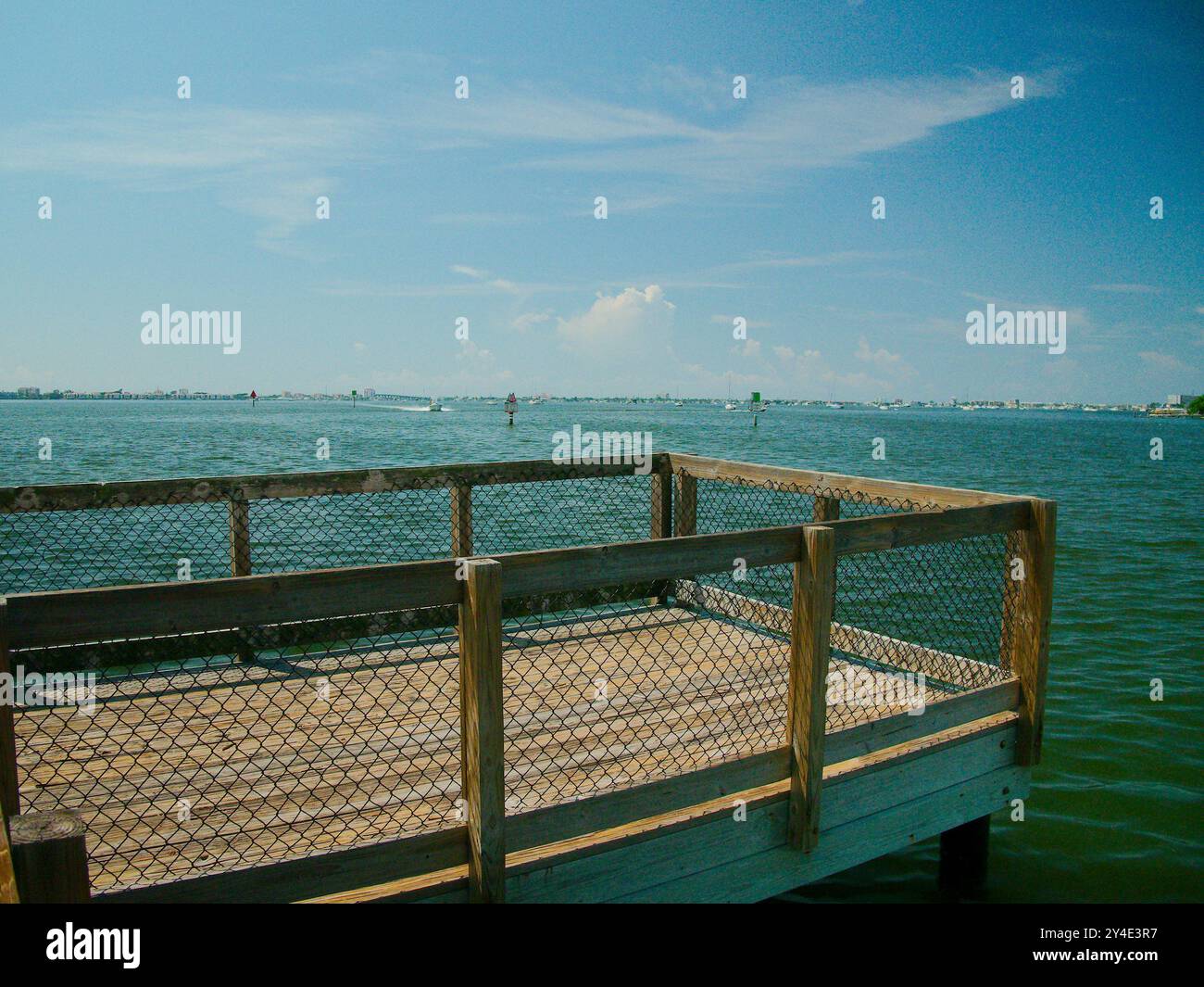 Weiter Blick über den Holzsteg Pier über die Boca Ciega Bay in Gulfport, FL. Hellblauer Himmel mit weißen Wolken und ruhigem Wasser. Boot nähert sich Stockfoto