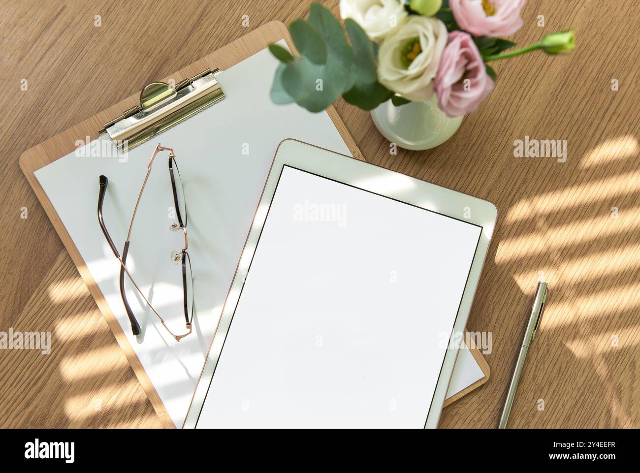 Ein heller Arbeitsbereich mit leerem Tablet, offenem Notizbuch, Schreibutensilien und frischen Blumen auf einem Holztisch lädt zu Kreativität und Kreativität ein Stockfoto