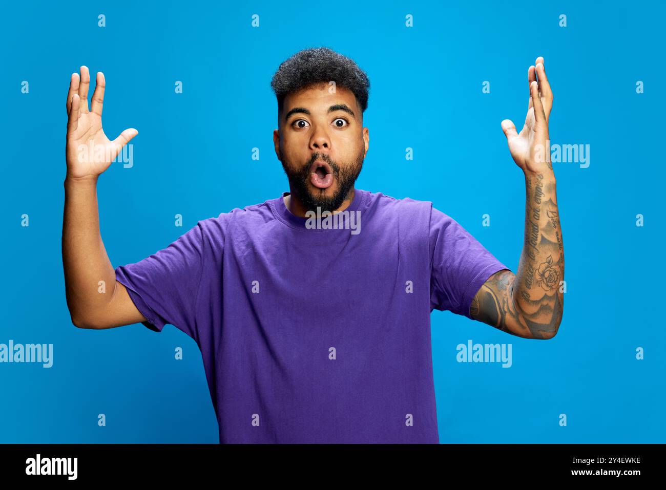 Ein afroamerikanischer Mann, der ein lila Hemd trägt, sieht überrascht aus, als er den Mund offen hält und seine Hände in verspielter Geste vor blauem Hintergrund hebt. Stockfoto