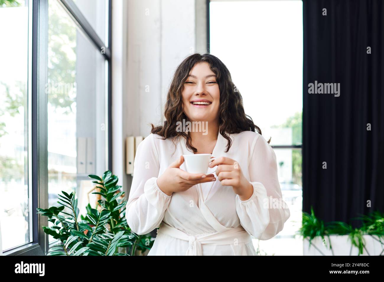 Eine junge Frau lächelt hell, während sie eine Tasse Kaffee in ihrem gemütlichen Büro hält. Stockfoto