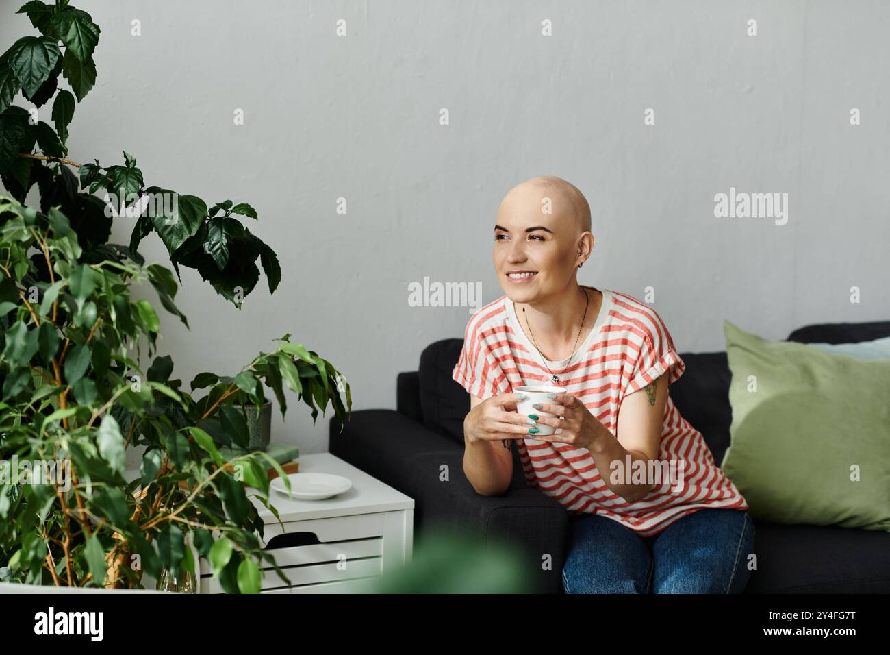 Eine fröhliche kahlköpfige Frau entspannt sich auf einer Couch, hält eine Tasse, inmitten üppiger Zimmerpflanzen und sanfter Einrichtung. Stockfoto