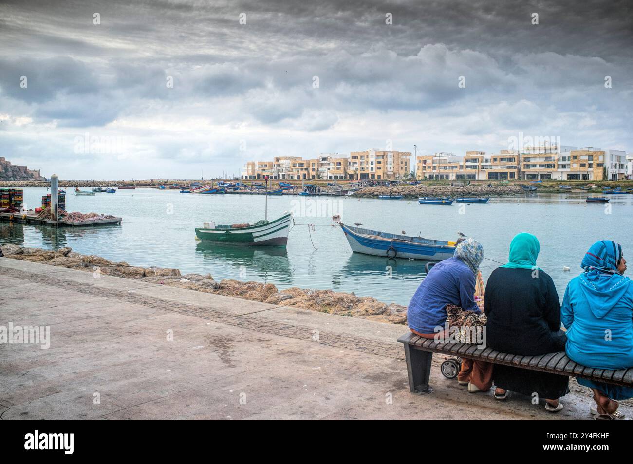 Die Menschen entspannen sich entlang des Flusses Bou Regreg in Rabat und genießen den Blick auf die Boote und die Stadt unter einem dramatischen bedeckten Himmel. Stockfoto