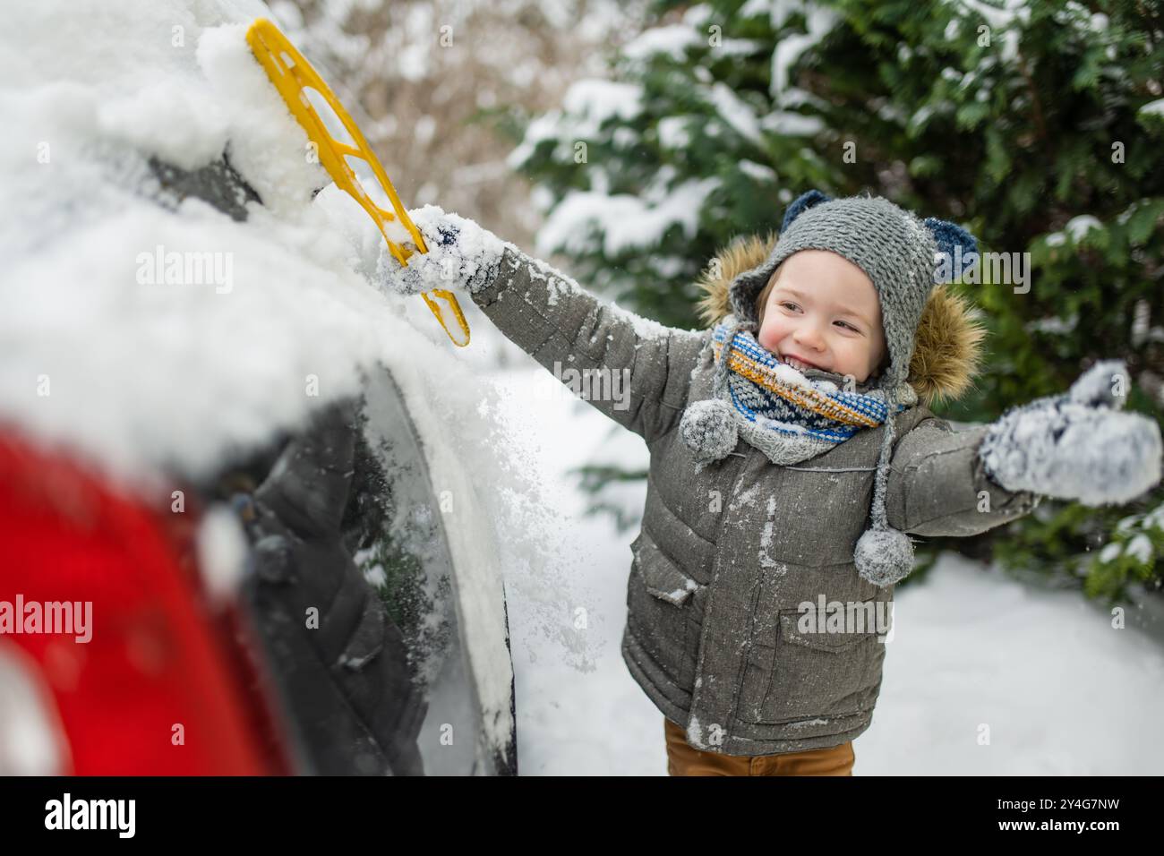 Ein entzückender kleiner Junge, der hilft, einen Schnee aus einem Auto zu bürsten. Mommy ist kleine Helferin. Winteraktivitäten für kleine Kinder. Stockfoto