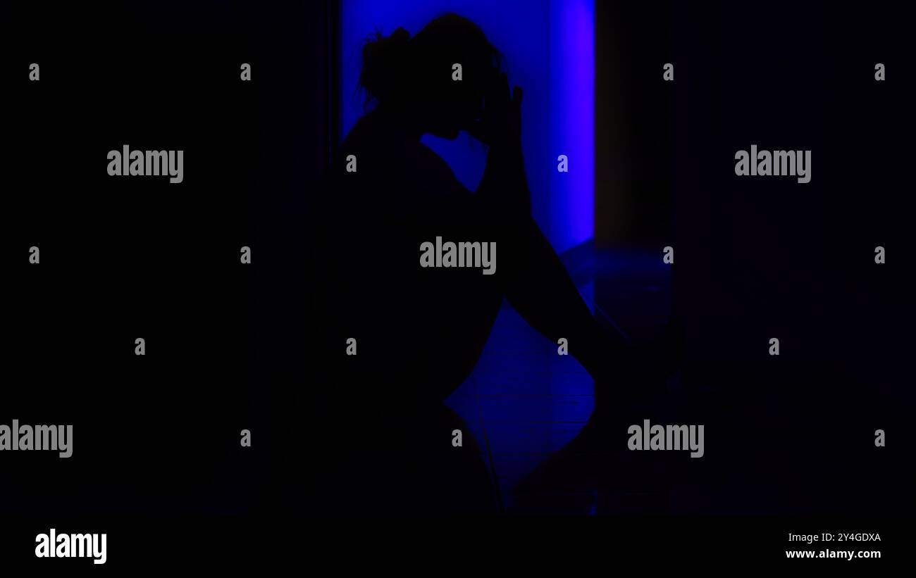 Eine Person sitzt allein in einem dunklen Flur, deren Silhouette von einem sanften blauen Licht beleuchtet wird, was ein Gefühl von Isolation und Introspektion vermittelt. Stockfoto