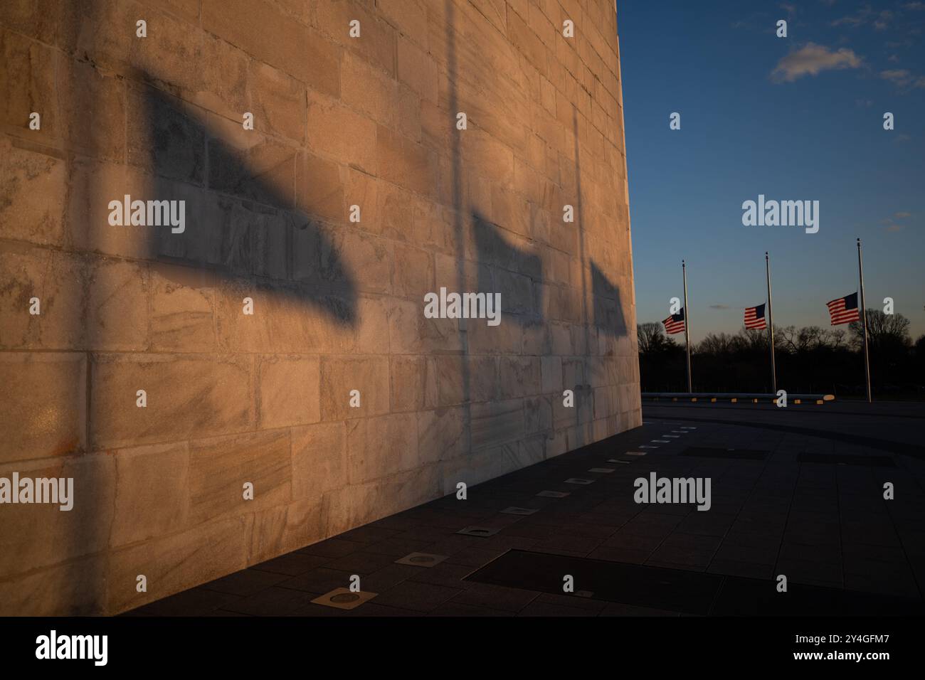 WASHINGTON Monument Flags at Sunset // WASHINGTON, Vereinigte Staaten – Schatten von amerikanischen Flaggen, die den Fuß des Washington Monuments umkreisen, erstrecken sich während des Sonnenuntergangs über seine Oberfläche. Der niedrige Winkel der Sonne erzeugt lange Silhouetten, die das Zusammenspiel zwischen dem Nationalsymbol und dem berühmten Obelisken in der National Mall betonen. Stockfoto