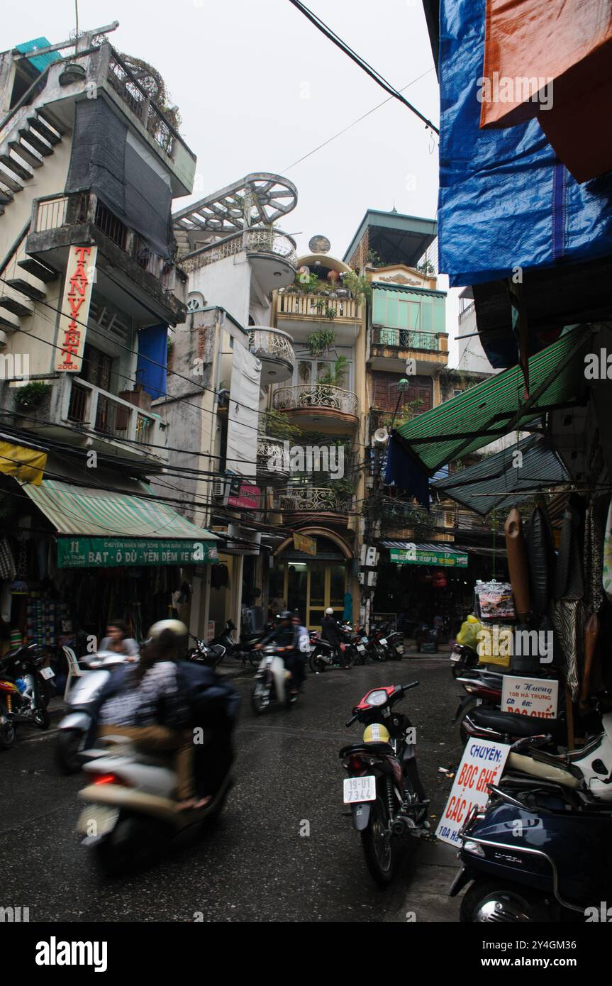 Straßenszene Motorräder Hanoi Vietnam // HANOI, Vietnam — Eine geschäftige Straßenszene in Hanoi zeigt alte Gebäude, die über einer engen, überfüllten Straße thronen. Die pulsierende Stadtlandschaft ist voller Motorräder, Straßenverkäufer und Fußgänger, die durch die lebhafte Atmosphäre der Stadt navigieren. Stockfoto