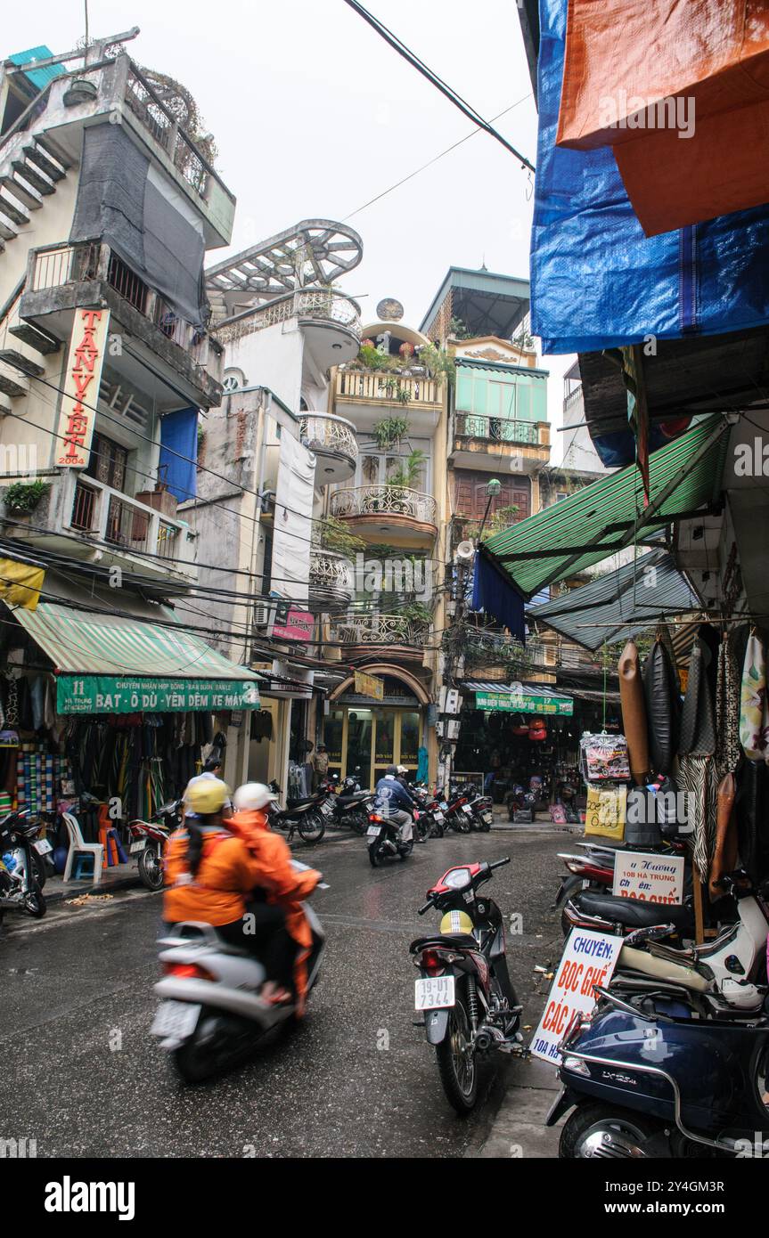 Straßenszene Motorräder Hanoi Vietnam // HANOI, Vietnam — Eine geschäftige Straßenszene in Hanoi zeigt alte Gebäude, die über einer engen, überfüllten Straße thronen. Die pulsierende Stadtlandschaft ist voller Motorräder, Straßenverkäufer und Fußgänger, die durch die lebhafte Atmosphäre der Stadt navigieren. Stockfoto