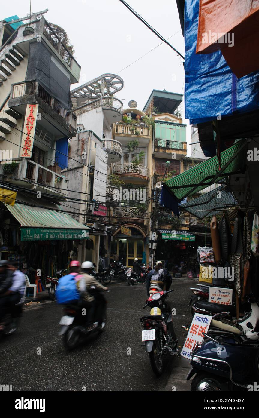 Straßenszene Motorräder Hanoi Vietnam // HANOI, Vietnam — Eine geschäftige Straßenszene in Hanoi zeigt alte Gebäude, die über einer engen, überfüllten Straße thronen. Die pulsierende Stadtlandschaft ist voller Motorräder, Straßenverkäufer und Fußgänger, die durch die lebhafte Atmosphäre der Stadt navigieren. Stockfoto