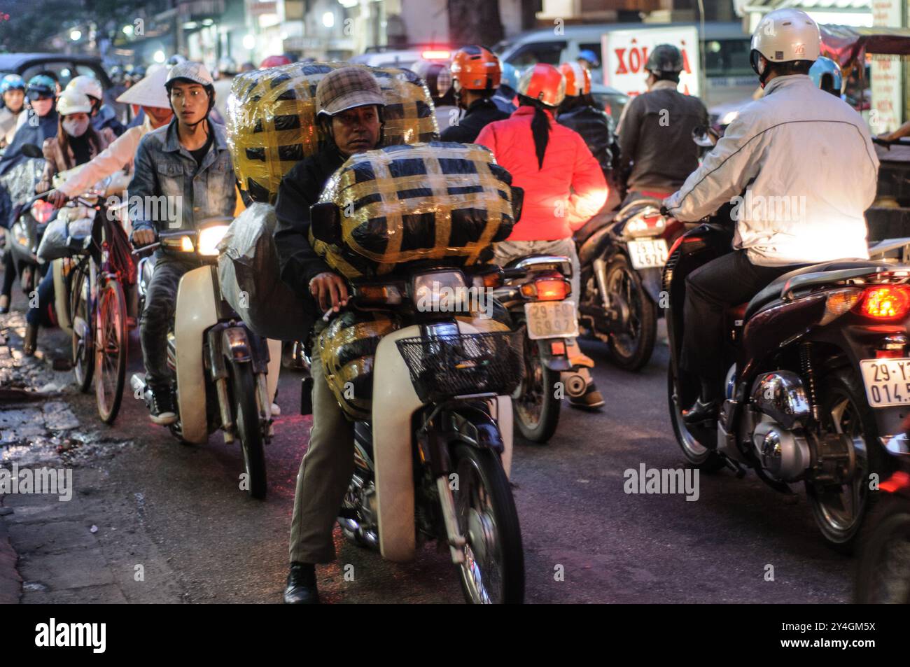 Motorroller und Fahrräder Hanoi Vietnam // HANOI, Vietnam – Motorroller und Fahrräder drängen sich am Abend auf eine enge Straße in der Altstadt von Hanoi. Die belebte Szene zeigt das chaotische, aber lebendige Straßenleben, das typisch für Hanoi ist, während die Einheimischen die geschäftigen Straßen mit dem Motorrad oder dem Fahrrad erkunden. Stockfoto