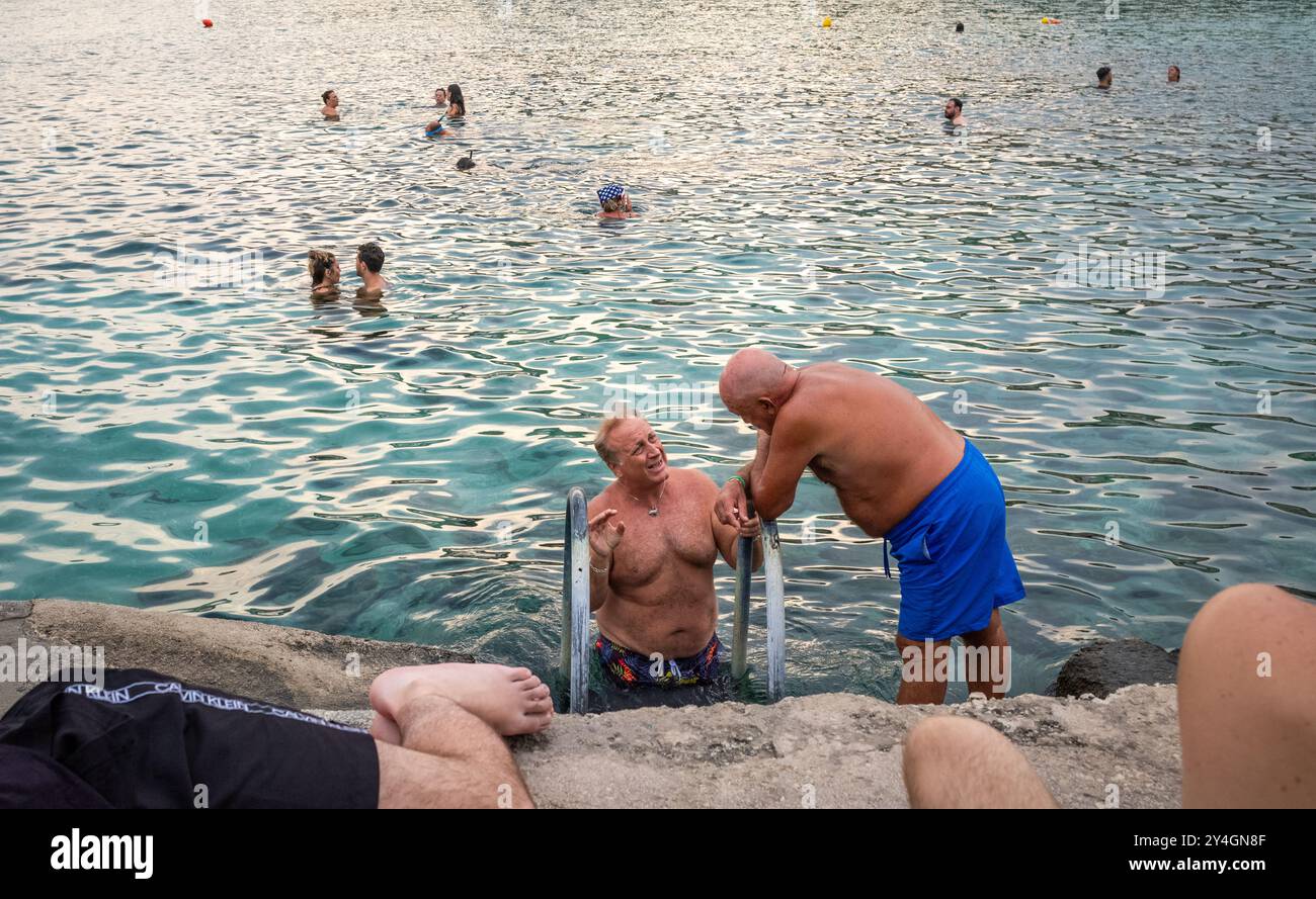 Zwei Männer sprechen, während die Italiener neben dem Wellenbrecher in Castro Marina, Apulien, Italien baden und schwimmen. Stockfoto