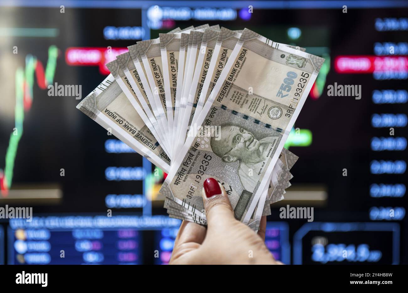 Eine Person, die eine Banknote in indischer Währung in 500 Rupien gegen ein Diagramm für den Aktienmarkt auf einem Display hält Stockfoto