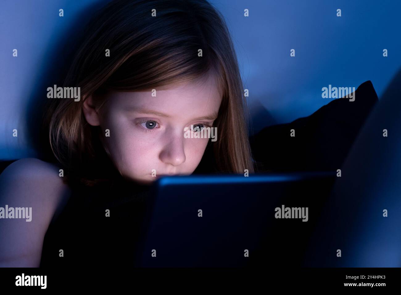 Porträt eines neunjährigen Mädchens bei schwachem Licht, das nachts auf einem Tablet-Bildschirm mit blauem Licht reflektiert. Stockfoto