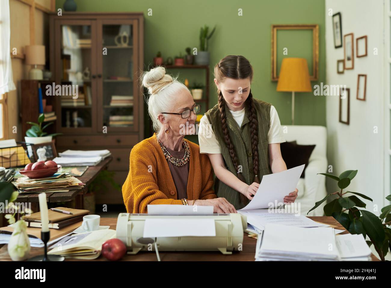 Junge Frau hilft älteren Frauen bei der Verwaltung von Dokumenten am Schreibtisch in einem warmen, einladenden Home Office-Ambiente mit Pflanzen und natürlichen Elementen, die eine friedliche Umgebung schaffen Stockfoto