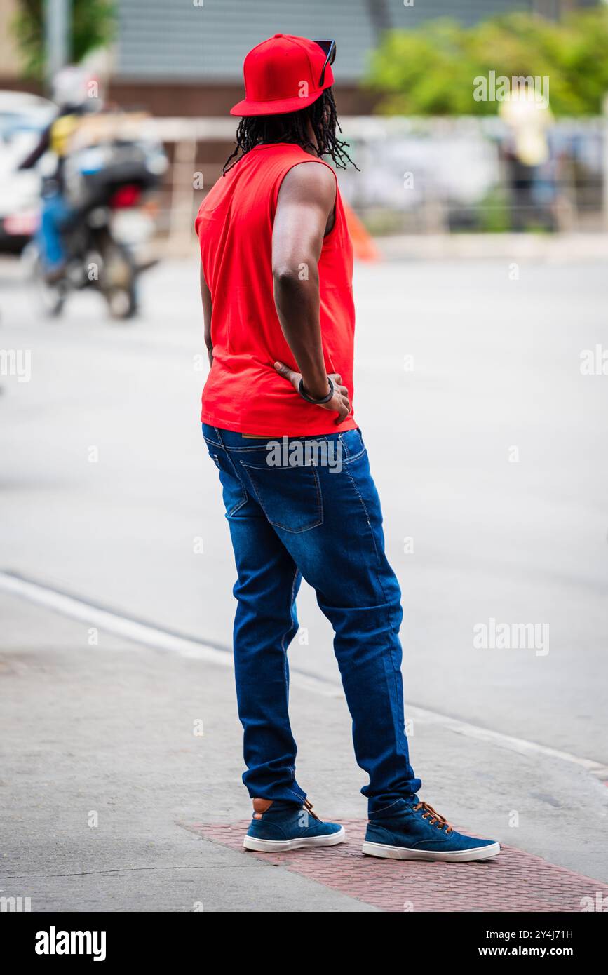 Schwarzer Mann mit rotem Hemd, rotem Hut und Jeans, der mit Hand auf Hüfte am Straßenrand in Belo Horizonte, Brasilien, steht. Stockfoto
