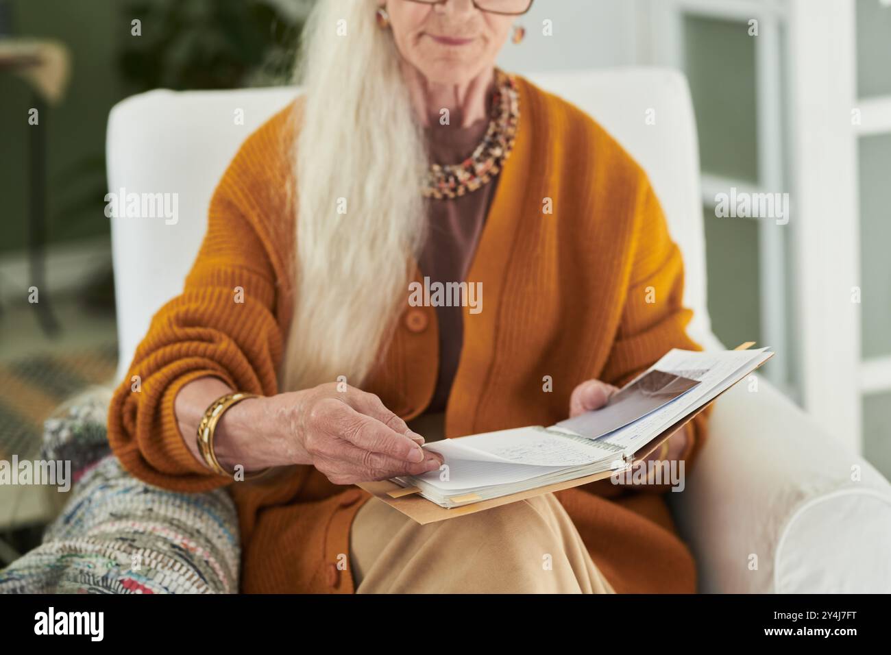 Ältere Frau mit langen grauen Haaren liest Buch, während sie bequem im Stuhl zu Hause sitzt und einen friedlichen und entspannten Moment zeigt Stockfoto