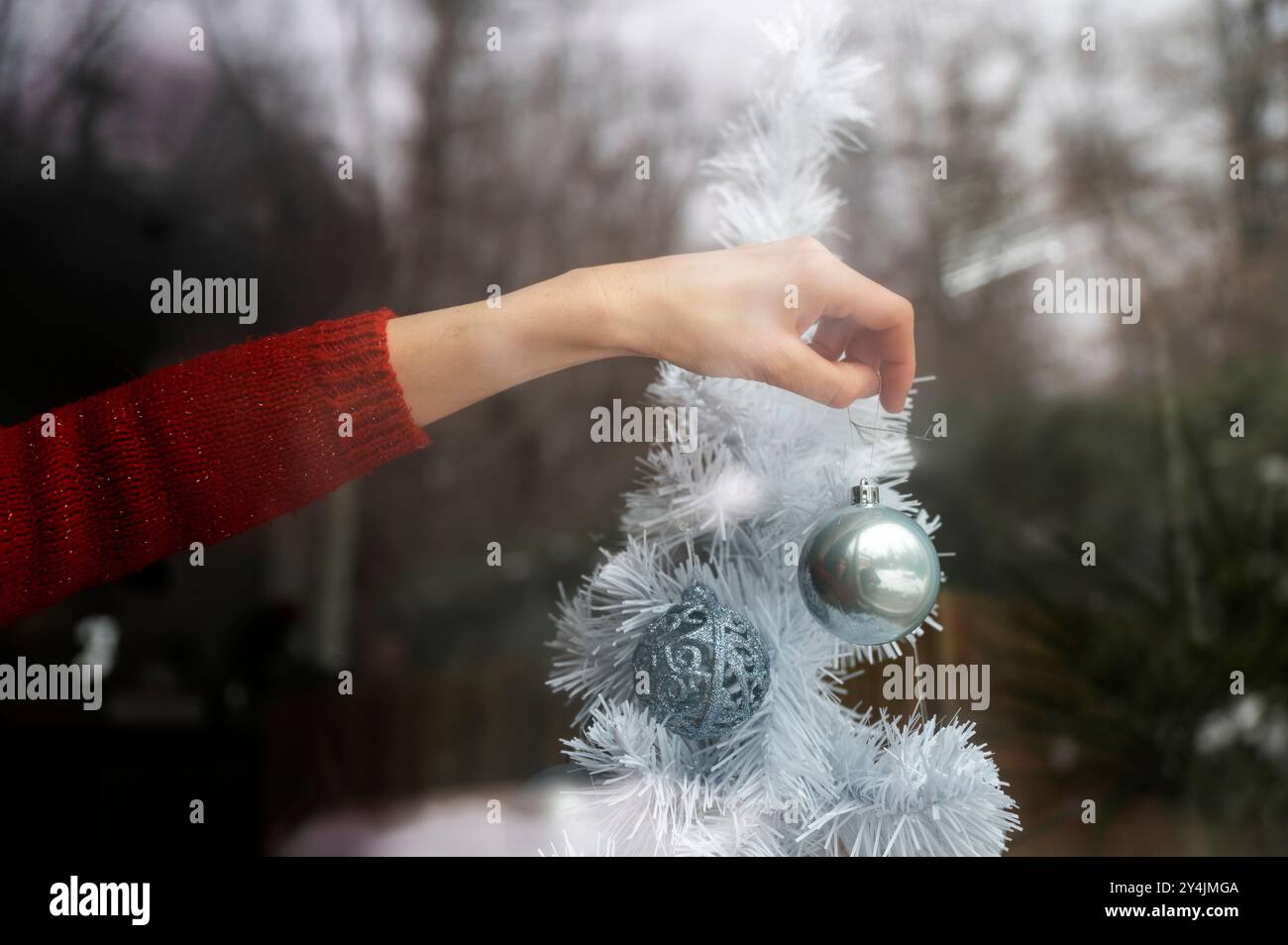 Blick durch das Fenster der weiblichen Hand in festlichem rotem Pullover, der glänzende blaue Weihnachtskugel auf weißen weihnachtsbaum mit Reflexion der Außenseite platziert Stockfoto