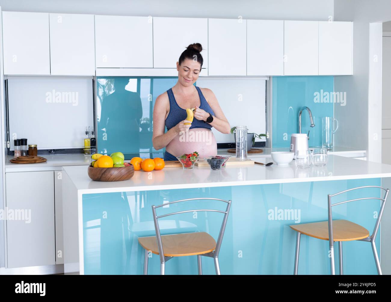 Zu Hause schält schwangere Frau Banane in moderner Küche mit frischem Obst auf der Theke Stockfoto