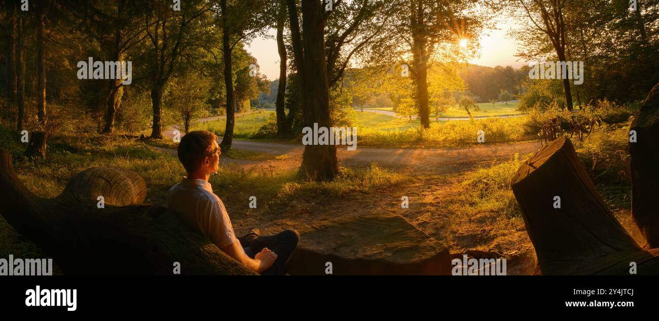 Ein junger Mann sitzt auf einer Bank in einem Park oder Wald und beobachtet die wunderschöne Landschaft, die von den Bäumen bei Sonnenuntergang eingerahmt wird Stockfoto