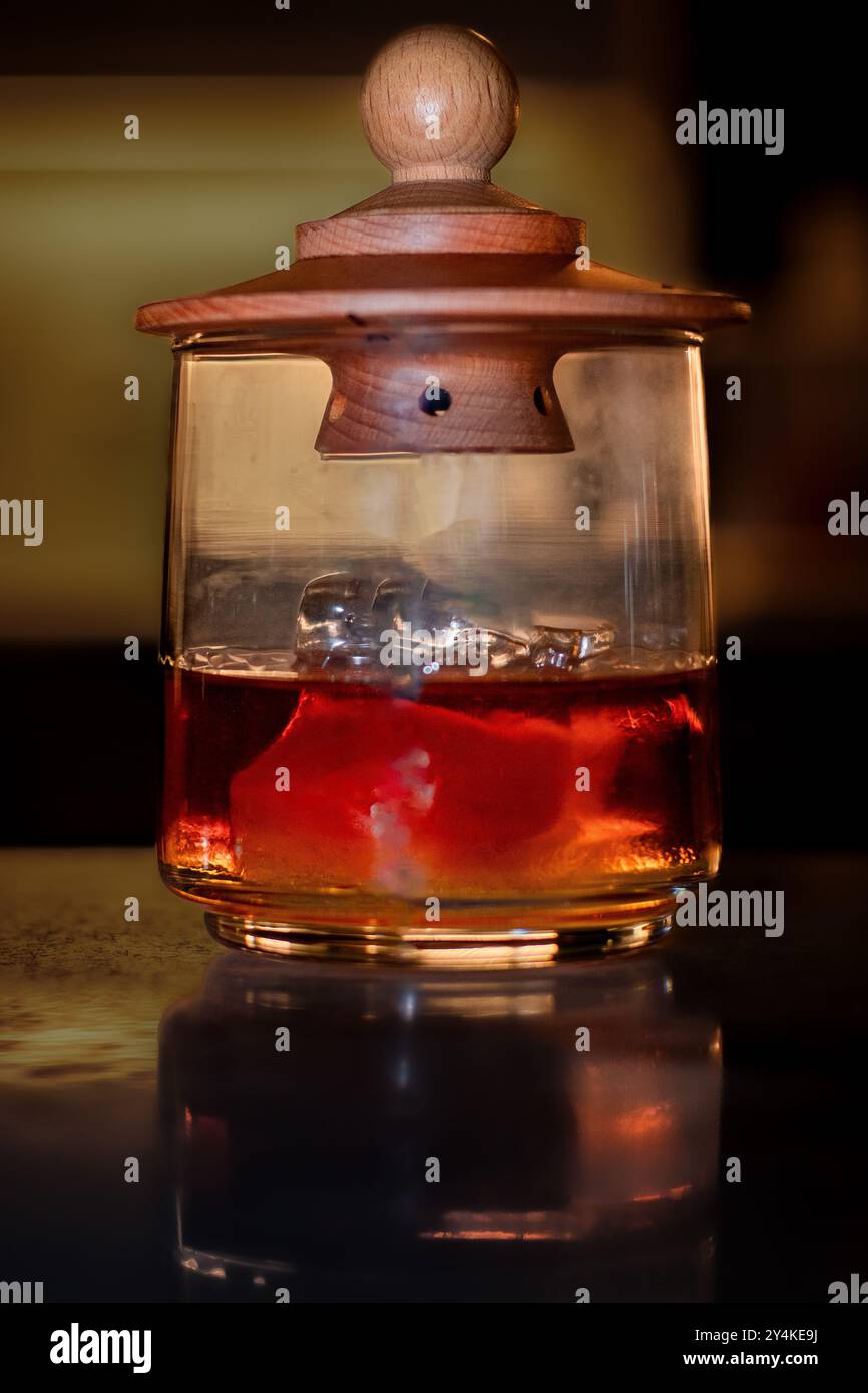 Ein geräucherter Whiskey-Cocktail in einem Glas mit Holzabdeckung. Die kräftige bernsteinfarbene Flüssigkeit wird vor einem dunklen Hintergrund beleuchtet, in der Innenseite ist ein subtiler Rauch sichtbar. Stockfoto