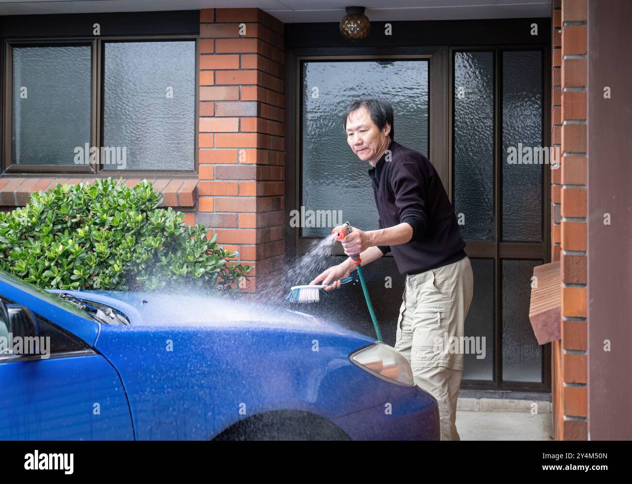 Ein Mann wäscht ein Auto zu Hause mit einer Bürste und einem Wasserspray aus einem Schlauch. Stockfoto