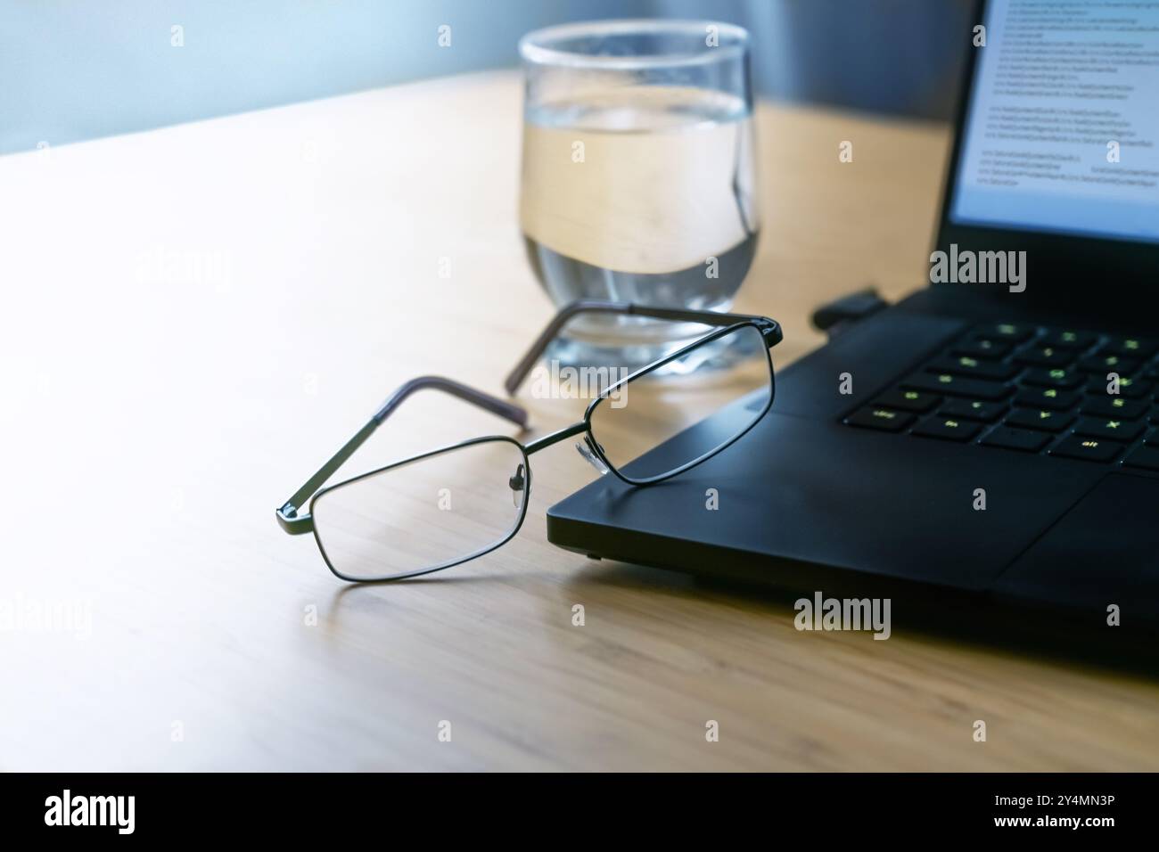 Brille, die sich gegen einen dunkelschwarzen Laptop mit Code auf einem Holzschreibtisch mit einem Glas Wasser im Büro lehnt, Geschäftskonzept bei kaltem Licht, Kopierraum, Stockfoto