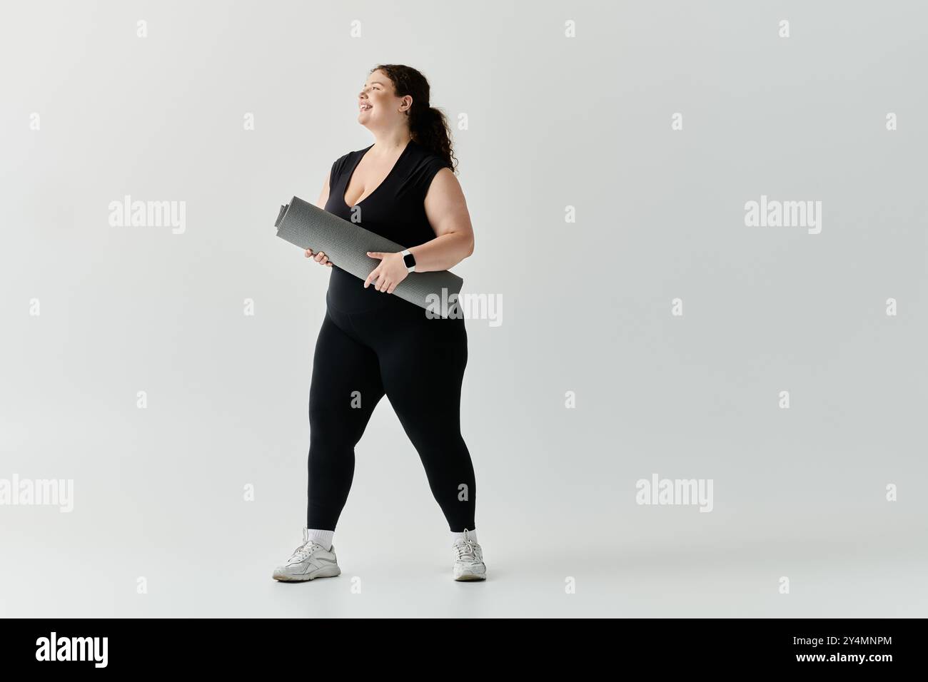 Eine elegante Frau in Übergröße steht selbstbewusst mit einer Yogamatte und lächelt in einem geräumigen Studio. Stockfoto