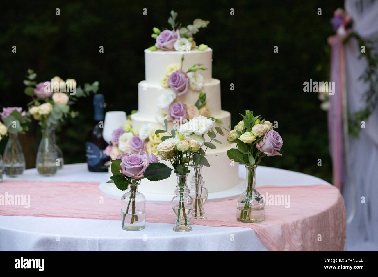 Weiße dreistufige Hochzeitstorte mit Rosen und Eustoma-Blumen, die auf einem Tisch mit rosa Tischdecke im Freien stehen Stockfoto