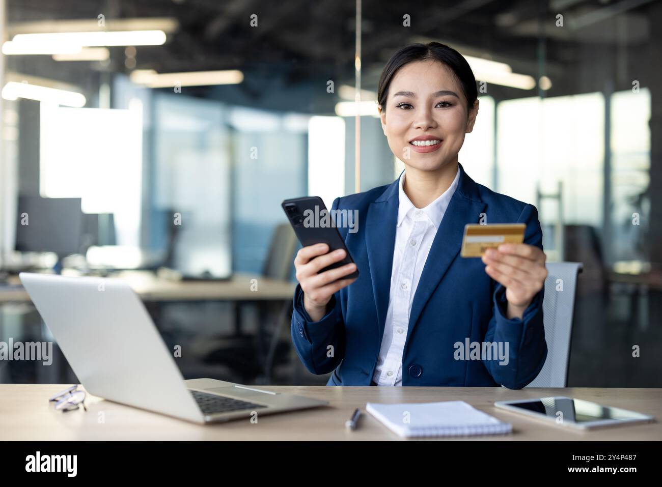Selbstbewusste asiatische Geschäftsfrau führt Online-Zahlungen per Smartphone und Kreditkarte im Büro durch. Zeigt professionelle Einstellung beim Multitasking mit Laptop und Telefon Stockfoto