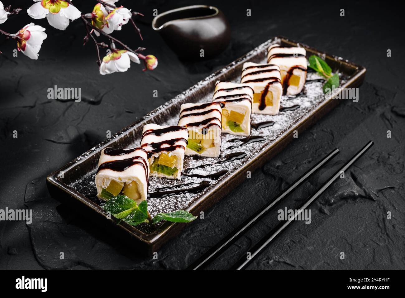 Elegante Dessert-Sushi-Auswahl mit Schokolade auf einem eleganten schwarzen Teller, geschmückt mit zarten Blumen Stockfoto