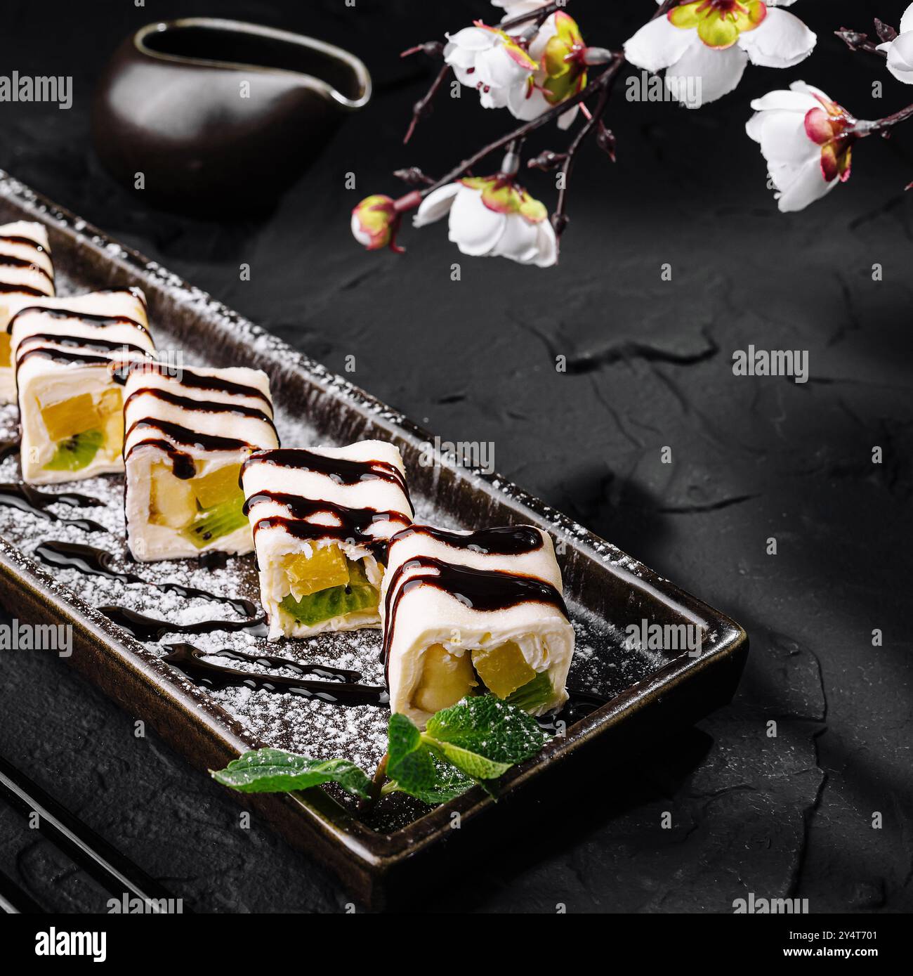 Elegante Dessert-Sushi-Auswahl mit Schokolade auf einem eleganten schwarzen Teller, geschmückt mit zarten Blumen Stockfoto